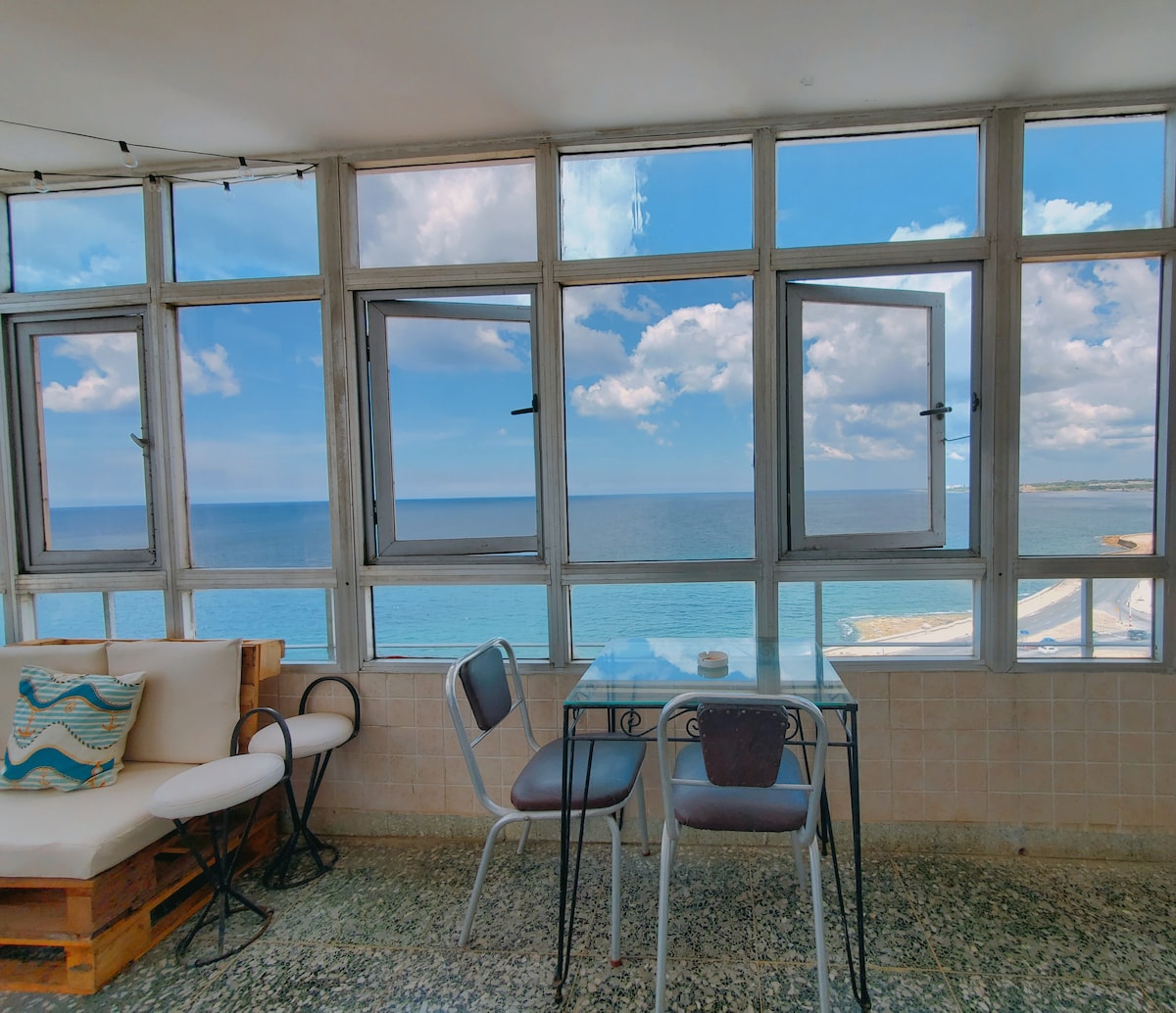 A bright seating area showcases large windows that offer expansive views of the sea and sky. A small dining table and chairs are positioned to enjoy the scenery. Comfortable seating in soft colors complements the inviting atmosphere, bathed in natural light.