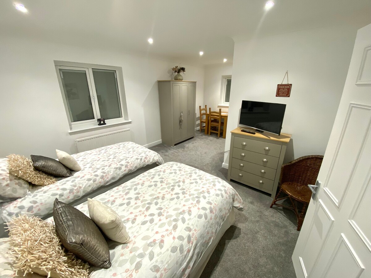 A well-furnished double bedroom features two single beds with light bedding and decorative pillows. A wardrobe and a dresser are visible, along with a TV mounted on the wall. A dining area is seen in the background, illuminated by soft overhead lighting.
