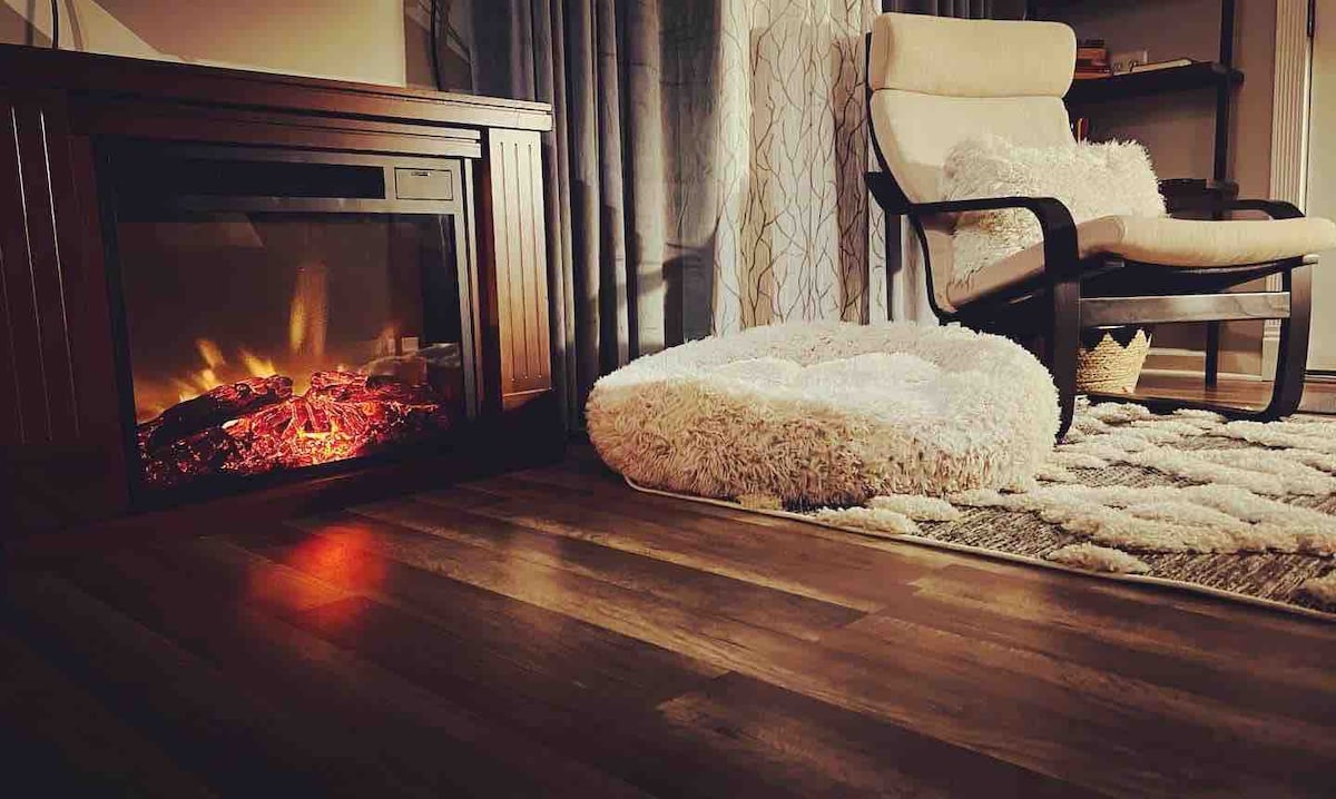 A cozy corner features a softly glowing electric fireplace and a plush dog bed on a textured area rug. A comfortable chair sits nearby, complemented by warm wood flooring and soft curtains, inviting moments of relaxation.