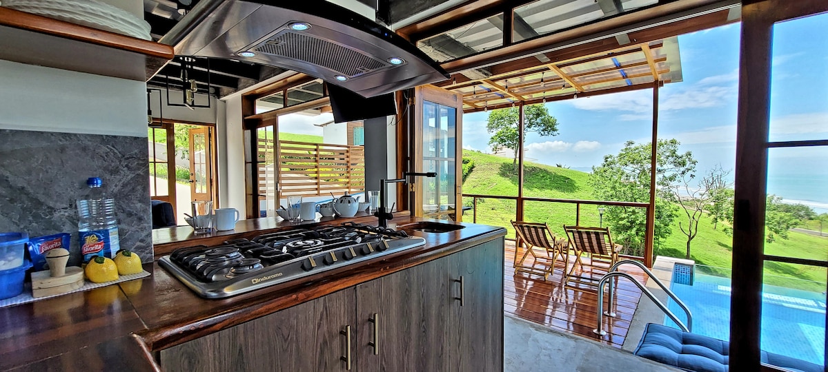A modern kitchen with a gas stove and sleek countertops opens to an outdoor deck and scenic ocean views. Natural light fills the space through large glass doors, creating a seamless connection to the infiniti pool and lush greenery beyond.