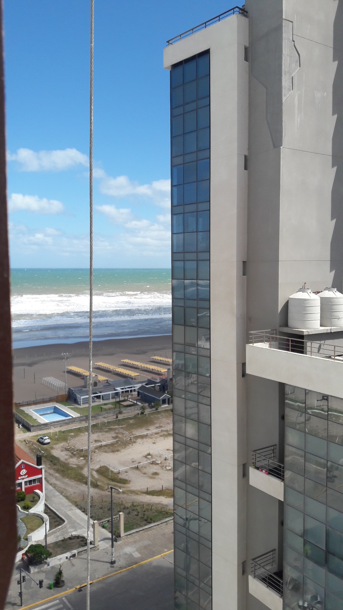 The image captures a view of a modern building alongside the coastline. The ocean is visible with waves gently rolling onto the sandy beach. A row of beach loungers can be seen, with a glimpse of a swimming pool in the distance. Clear blue skies are interspersed with scattered clouds.