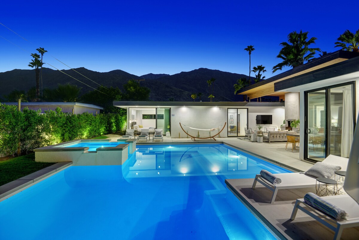 A stunning outdoor space is highlighted by a saltwater pool, reflecting the evening sky. Lounge chairs are positioned around the pool, alongside a hammock for relaxation. The modern home features large sliding glass doors that open to the pool area, with lush greenery providing privacy.