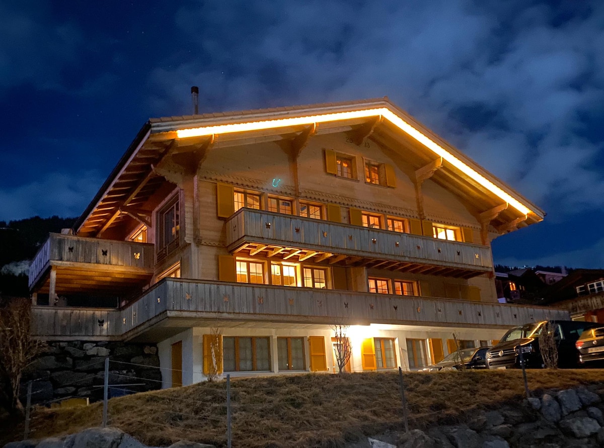 The chalet is presented at night, featuring a rustic wooden exterior with multiple balconies illuminated by warm lights. The spacious structure showcases large windows, offering glimpses of the interior. The surrounding area includes a grassy yard and parked vehicles.