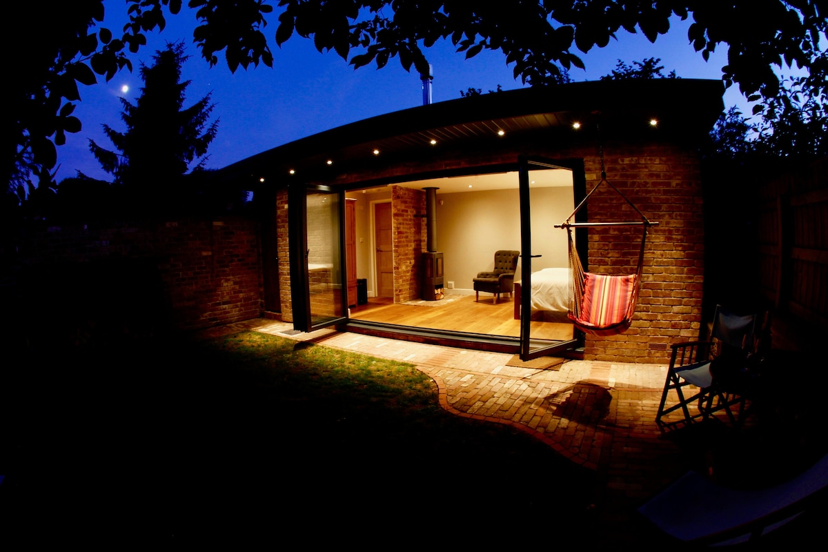 The garden studio is shown at dusk, featuring large bi-fold doors that open to a private lawn area. A hanging chair, illuminated by soft lighting, is suspended under an overhang. The wooden floor and cozy seating area create an inviting atmosphere.