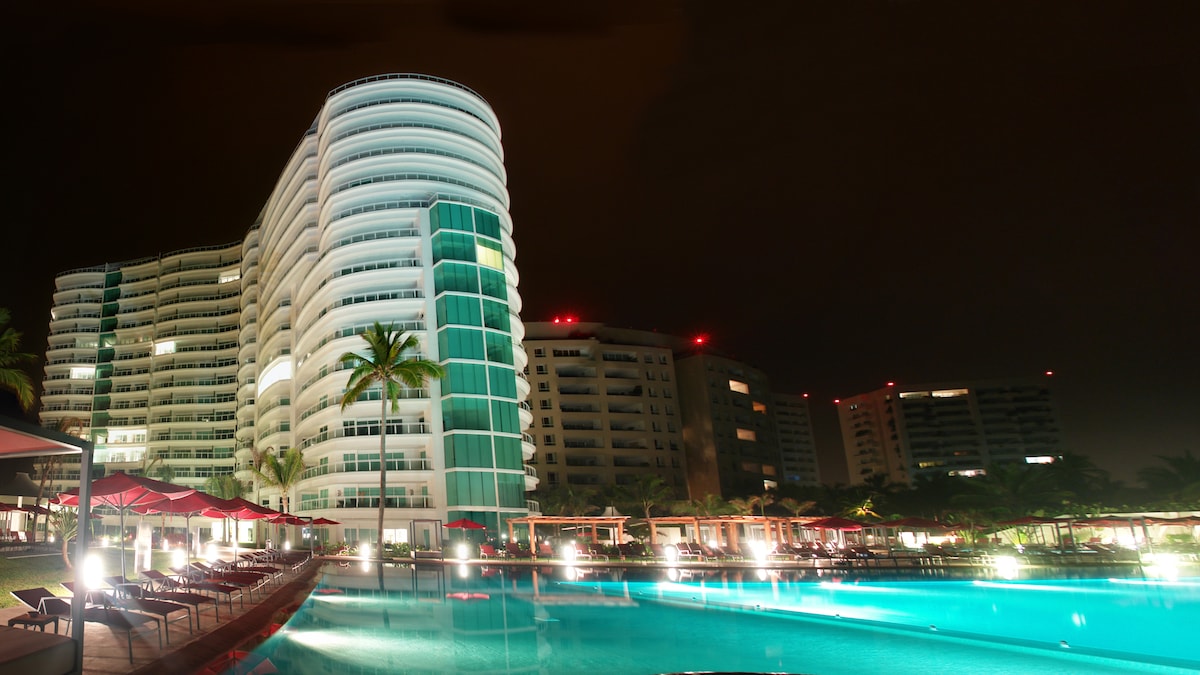 The image captures a tall, modern building illuminated against the night sky, with large windows reflecting the pool’s gentle blue light. Surrounding the pool, lounge chairs and palm trees are present, creating a serene resort atmosphere.