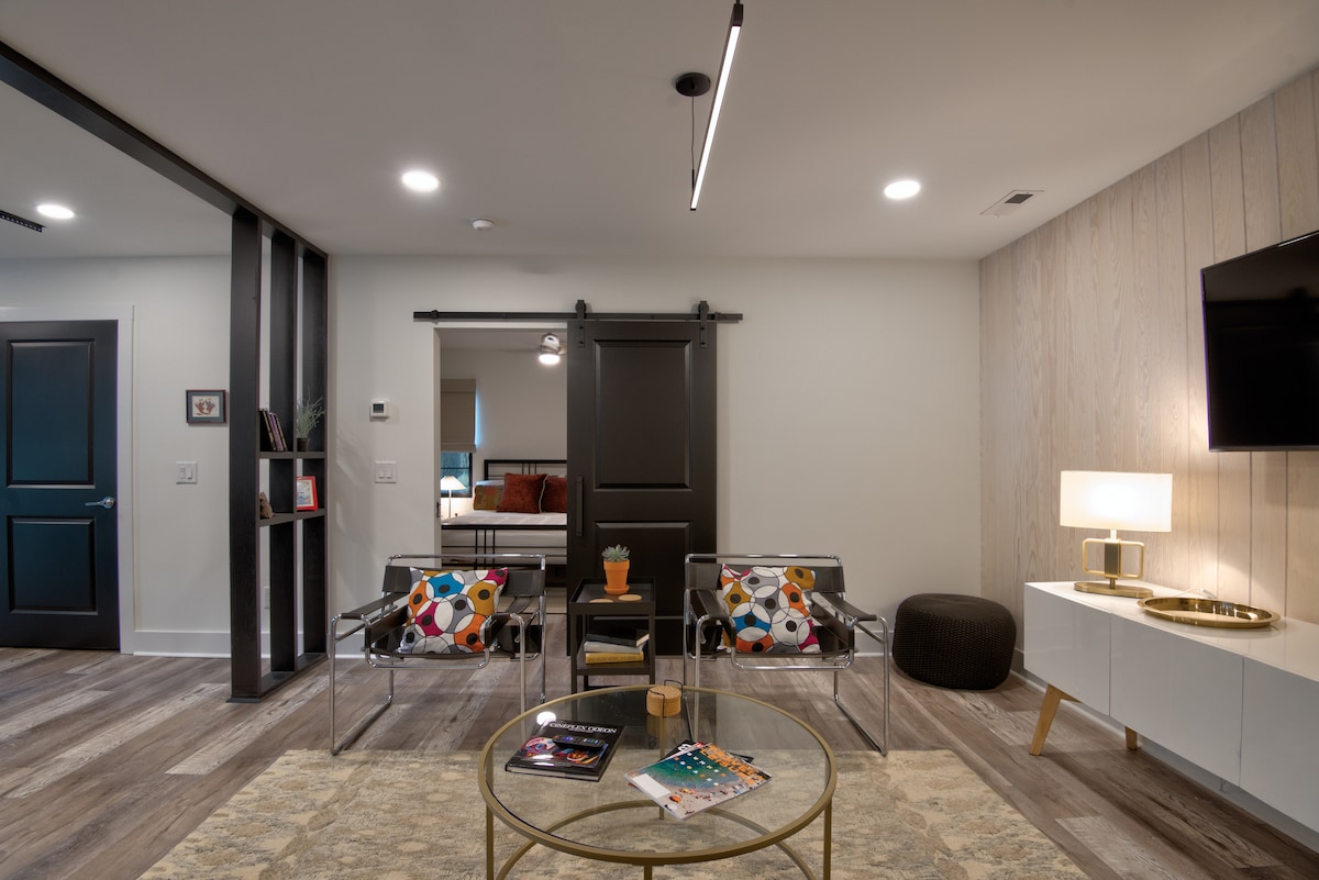 The living space features modern design elements, including a round coffee table and two lounge chairs with vibrant patterns. A large flat-screen TV is mounted on the wall, while natural light is provided through a sliding door and nearby windows, enhancing the welcoming ambiance.