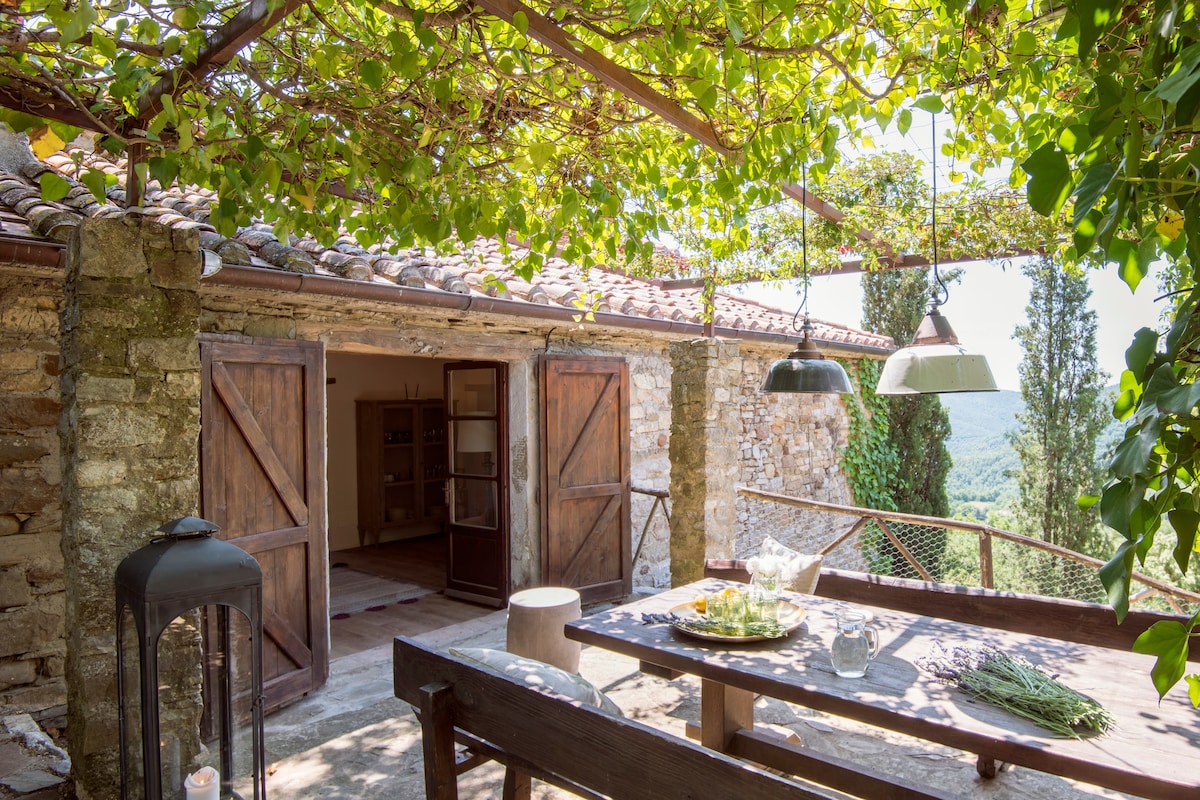 An outdoor dining area is set under a canopy of greenery, featuring a wooden table ready for a meal. Two hanging light fixtures provide illumination, while rustic stone walls surround the space. Open doors lead to an interior area with simple furnishings.