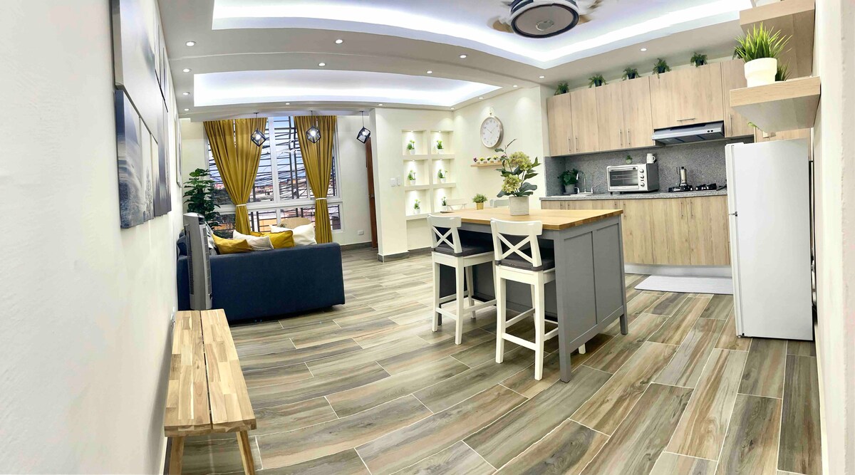 A light-filled living space features an open kitchen area with wooden cabinetry and modern appliances. The lounge is furnished with a dark sofa and a wooden bench. Bright yellow curtains frame the large window, complementing the warm tones of the tiled flooring.