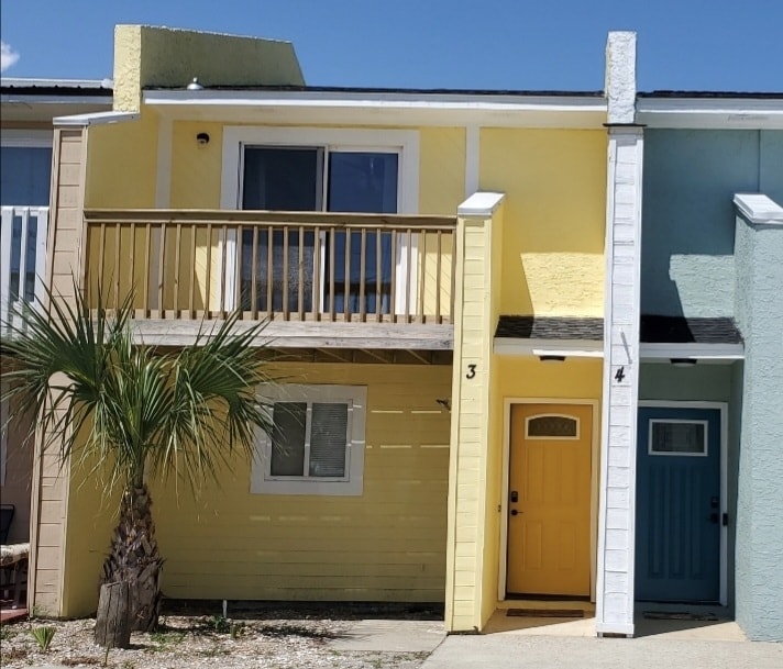 A two-story beach house is displayed, featuring a yellow exterior with a front porch and a bright blue door. A palm tree is visible in front, complementing the tropical setting. Light shades brighten the facade under a clear blue sky.