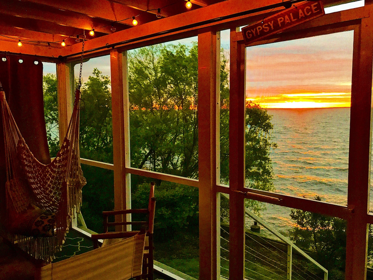 A sunlit sunroom provides expansive views of the Chesapeake Bay, framed by large glass windows. A hammock and a wooden chair are positioned for relaxation, with soft golden hues from the sunset reflecting on the water and trees just outside.