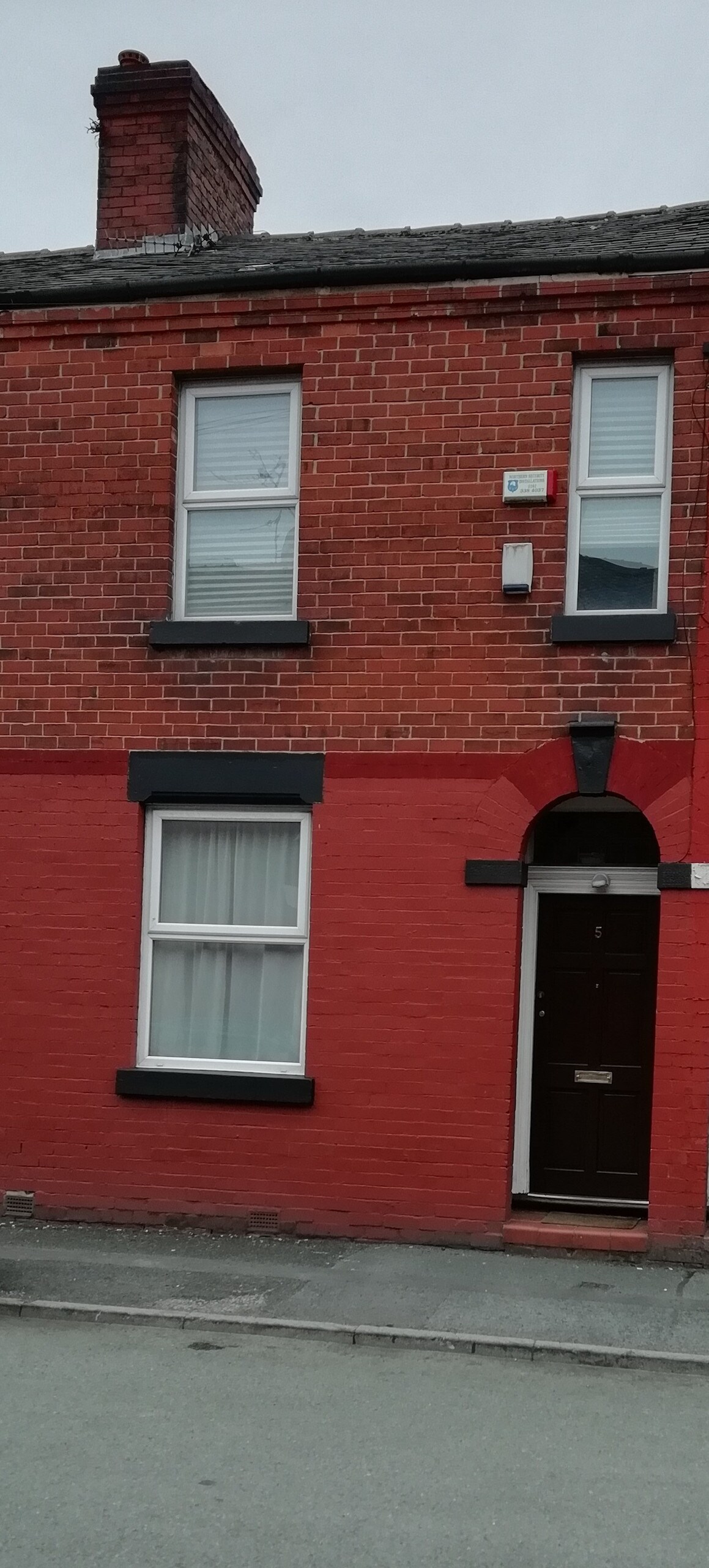 The exterior of a terraced house features a red-brick façade with black trim. Large windows allow natural light to enter, and a brown front door is centered within the structure. The house presents a neat and well-maintained appearance in a quiet street.