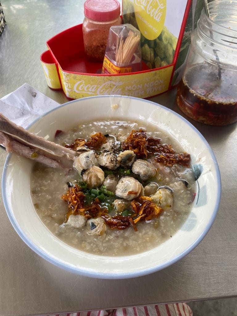 A bowl of rice porridge is presented, topped with various ingredients such as herbs and fried shallots. A side of dipping sauce, along with condiments, is visible in the background. A spoon with a piece of meat rests in the bowl, adding to the meal's visual appeal.