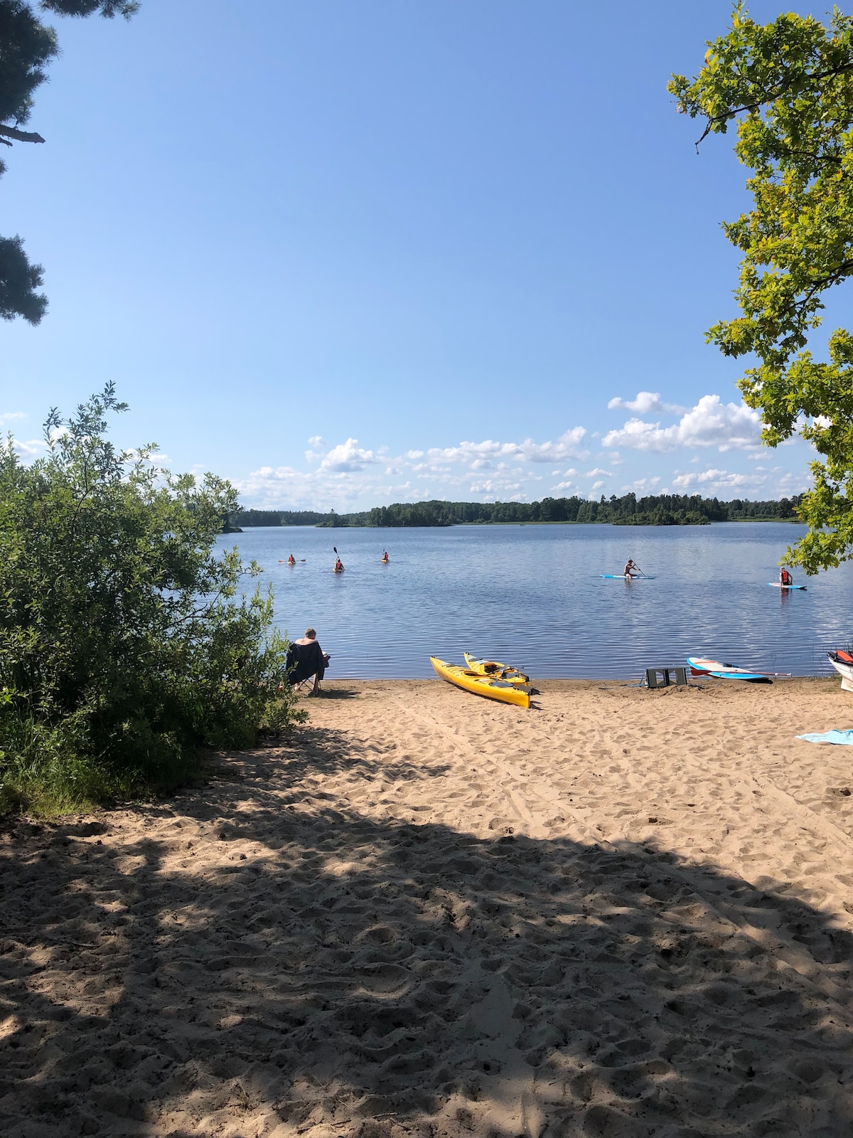 A sunny beach scene along Agunnaryd lake shows individuals kayaking and swimming in the water. Soft sand leads to the lake's edge, framed by green foliage on one side. Colorful kayaks are visible on the sand, inviting water activities in a serene setting.