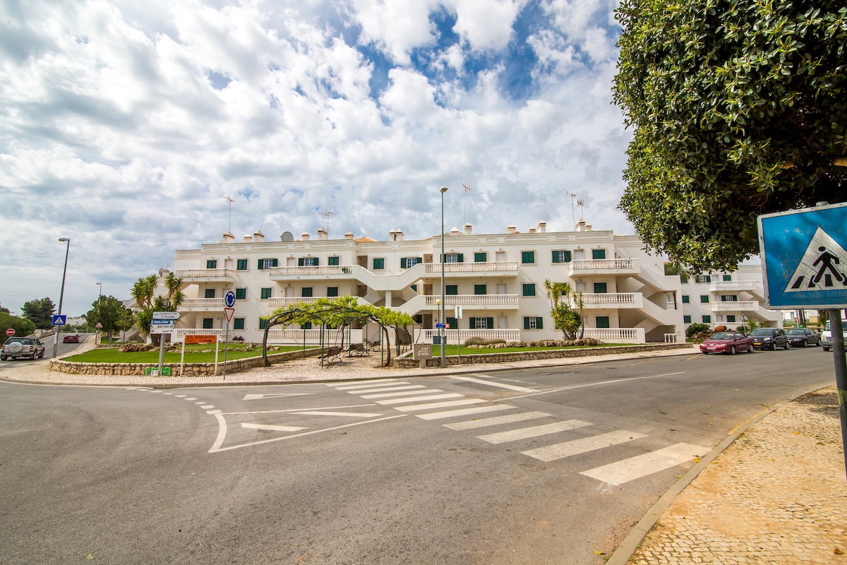 The apartment building features a modern architecture with multiple levels, accentuated by large windows and balconies. Greenery is visible around the property, enhancing the serene atmosphere. A crosswalk and road signage are present, indicating accessibility to the surrounding area.