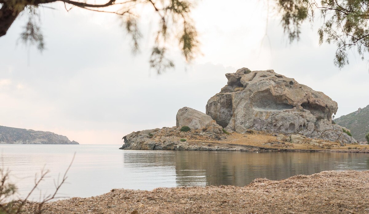 A rocky formation is visible along the shore, partially reflected in the calm waters. The surrounding landscape features gentle hills and sparse vegetation, while the sky above holds soft, diffused light, suggesting a serene atmosphere.
