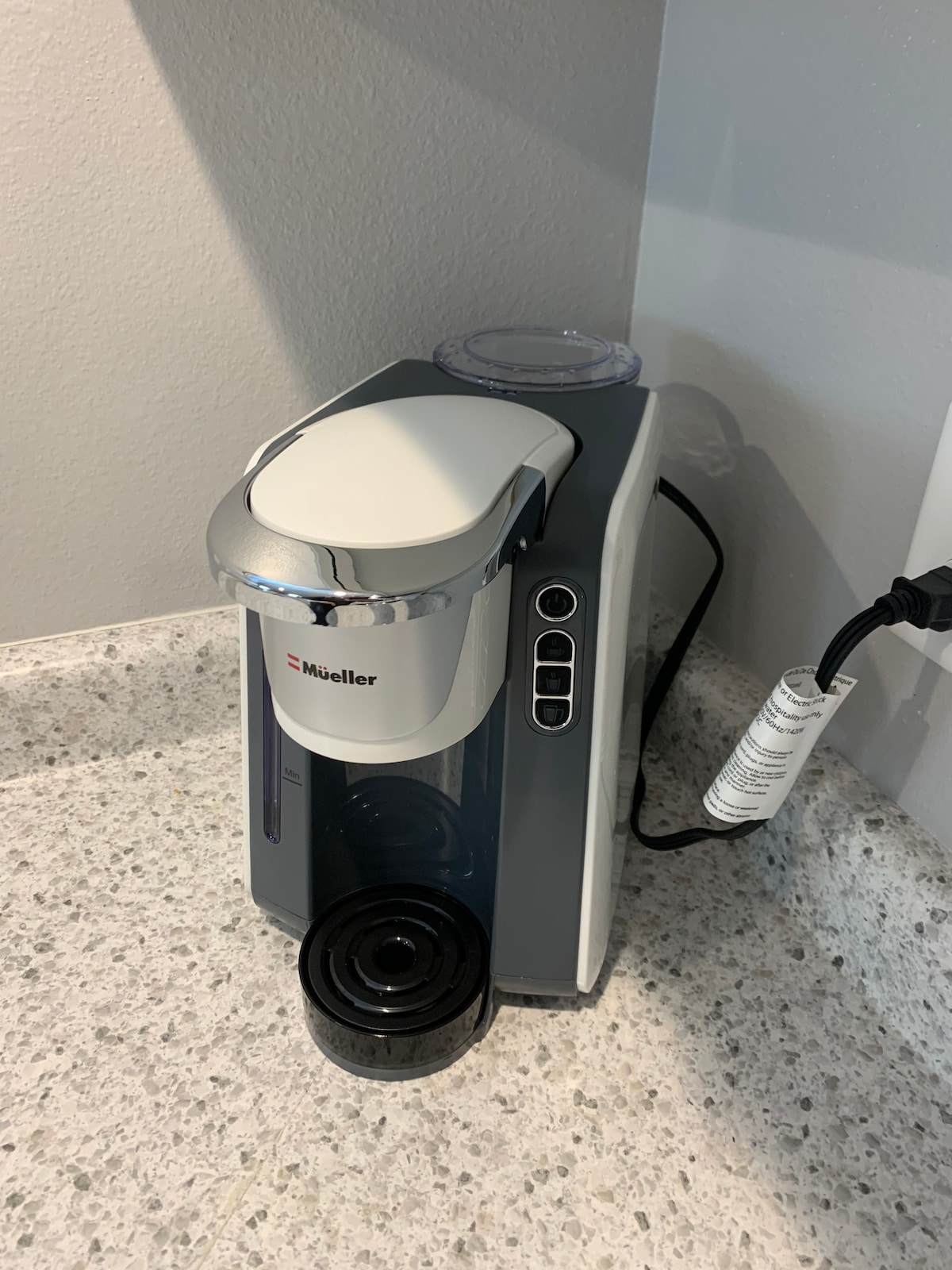 A modern coffee maker is positioned on a speckled countertop. The machine features a sleek design with a chrome finish and a visible water reservoir. A black coffee cup sits beneath the spout, ready for use.