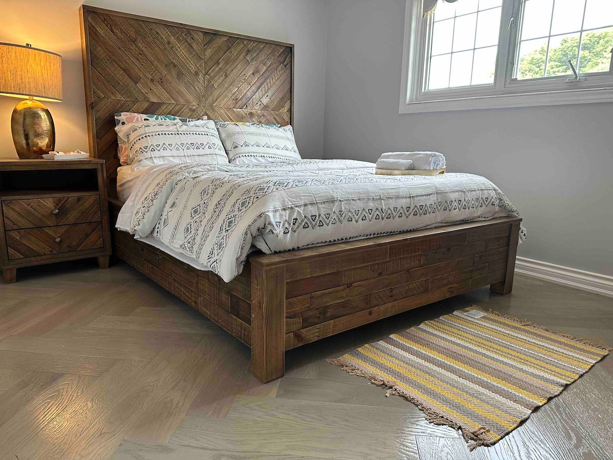 A cozy bedroom features a rustic wooden bed with a detailed headboard, dressed in soft, patterned linens. A nightstand with a lamp stands beside the bed, providing gentle illumination. A striped rug adds texture to the hardwood floor, while natural light filters through the window.