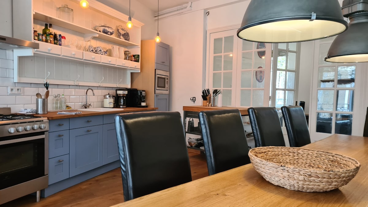 An open kitchen and dining area features a large wooden table with several black leather chairs. Shelves display dishware, and modern appliances are integrated in the light blue cabinetry. Warm pendant lights hang above, creating a functional and inviting cooking space.