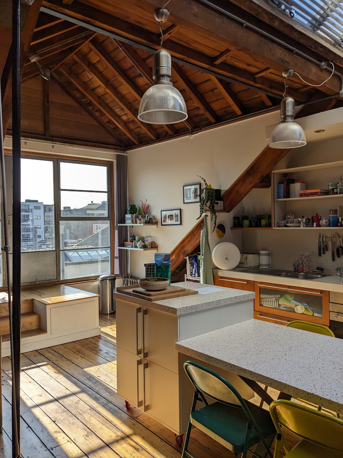 A bright kitchen area features high wooden ceilings with exposed beams, and large windows allowing natural light to fill the space. A central island with a countertop is complemented by open shelving, while eclectic seating options add a touch of color.