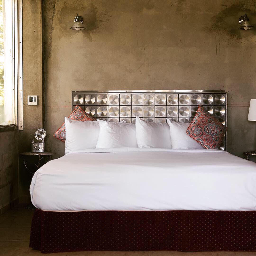 The bedroom features a spacious bed with a plush white duvet and decorative pillows. A textured metallic headboard catches the light. Natural light enters through a large window, enhancing the room's simplicity, while a stylish bedside table and lamp provide functional decor.