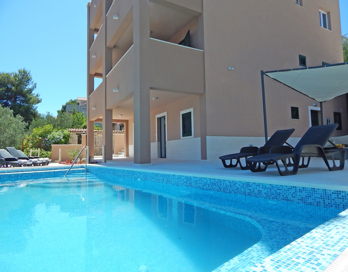 A modern building is displayed beside a clear swimming pool. Comfortable lounge chairs are positioned around the pool area, providing space for relaxation. A shaded seating area is visible in the background, surrounded by greenery, enhancing the outdoor experience.
