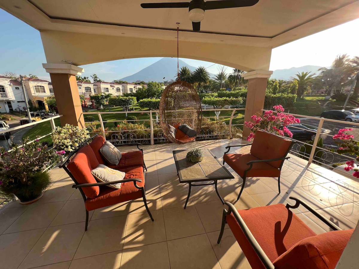 A balcony area features a hanging chair alongside two armchairs, each complemented by decorative cushions. A small table is positioned between the seating. Lush greenery and flowering plants frame the space, while a view of the Chaparrastique volcano is visible in the background.