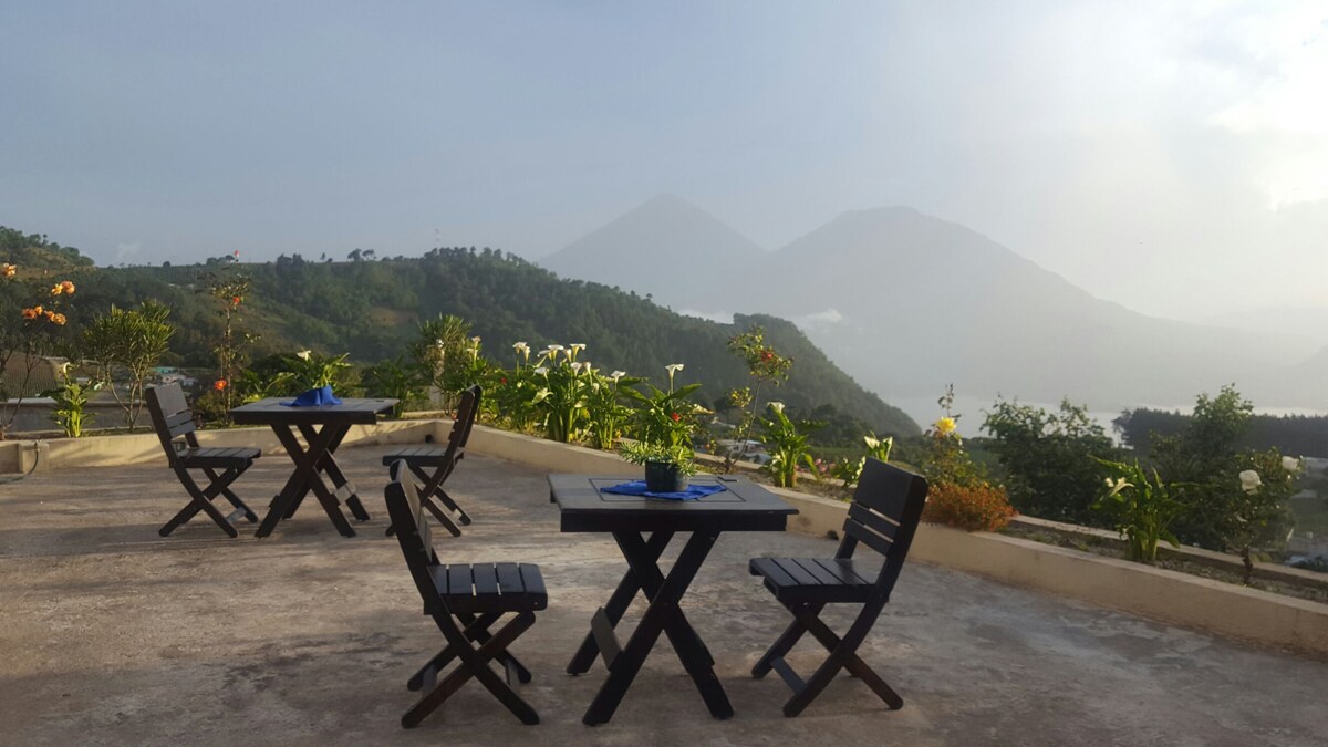 An outdoor terrace is furnished with four black wooden chairs and two tables, offering views of distant mountains and lush greenery. Flowering plants add color to the surroundings, creating a serene environment for relaxation and connection with nature.