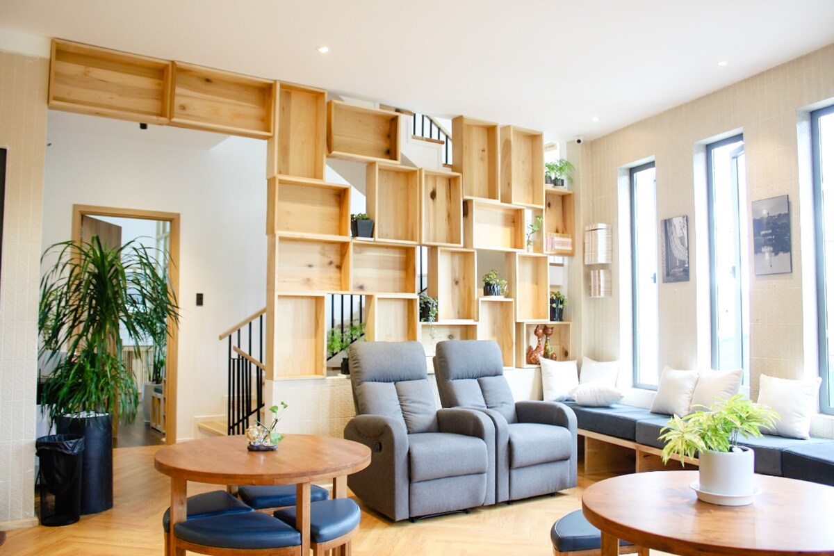 A contemporary lounge area features a combination of comfortable gray reclining chairs and wooden tables. A decorative shelving unit, filled with greenery and personal items, serves as a focal point. Large windows allow natural light to brighten the space.