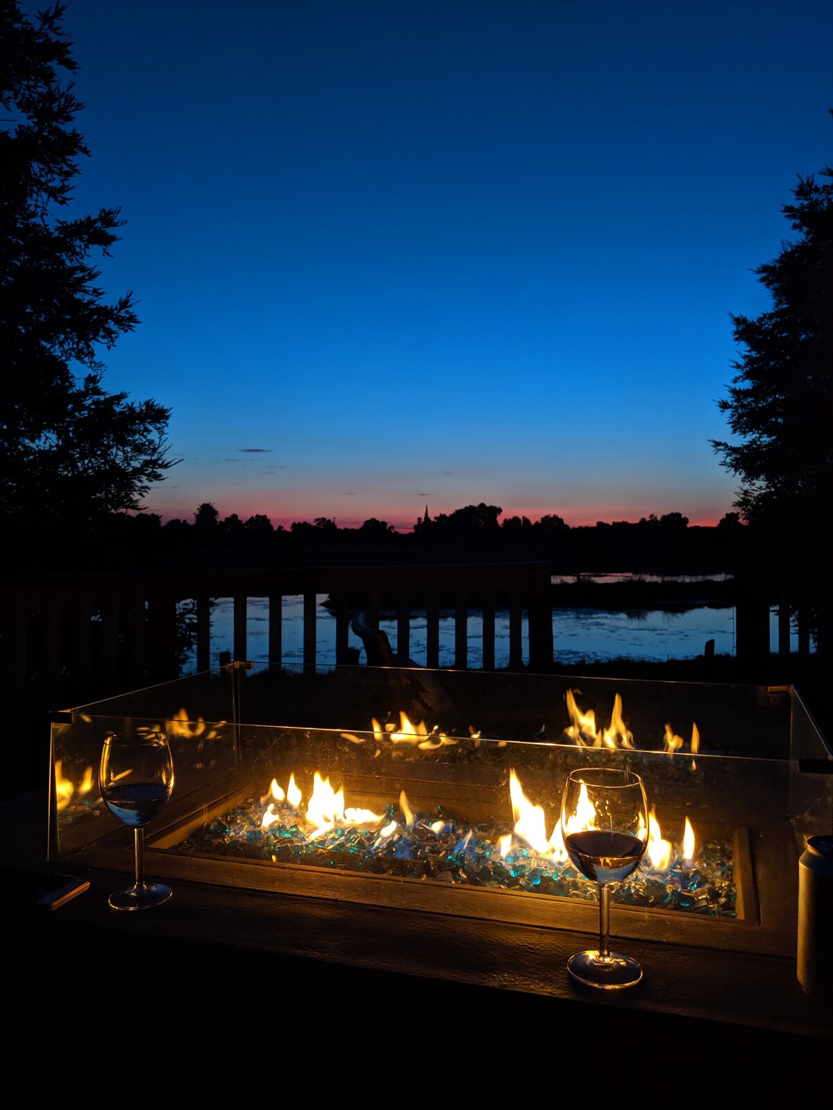 A fire pit with a glass enclosure glows brightly, casting warm light over the surrounding area. Two wine glasses are placed on the table, enhancing the serene setting. The tranquil water reflects the fading light of a colorful sunset sky filled with shades of blue and orange.