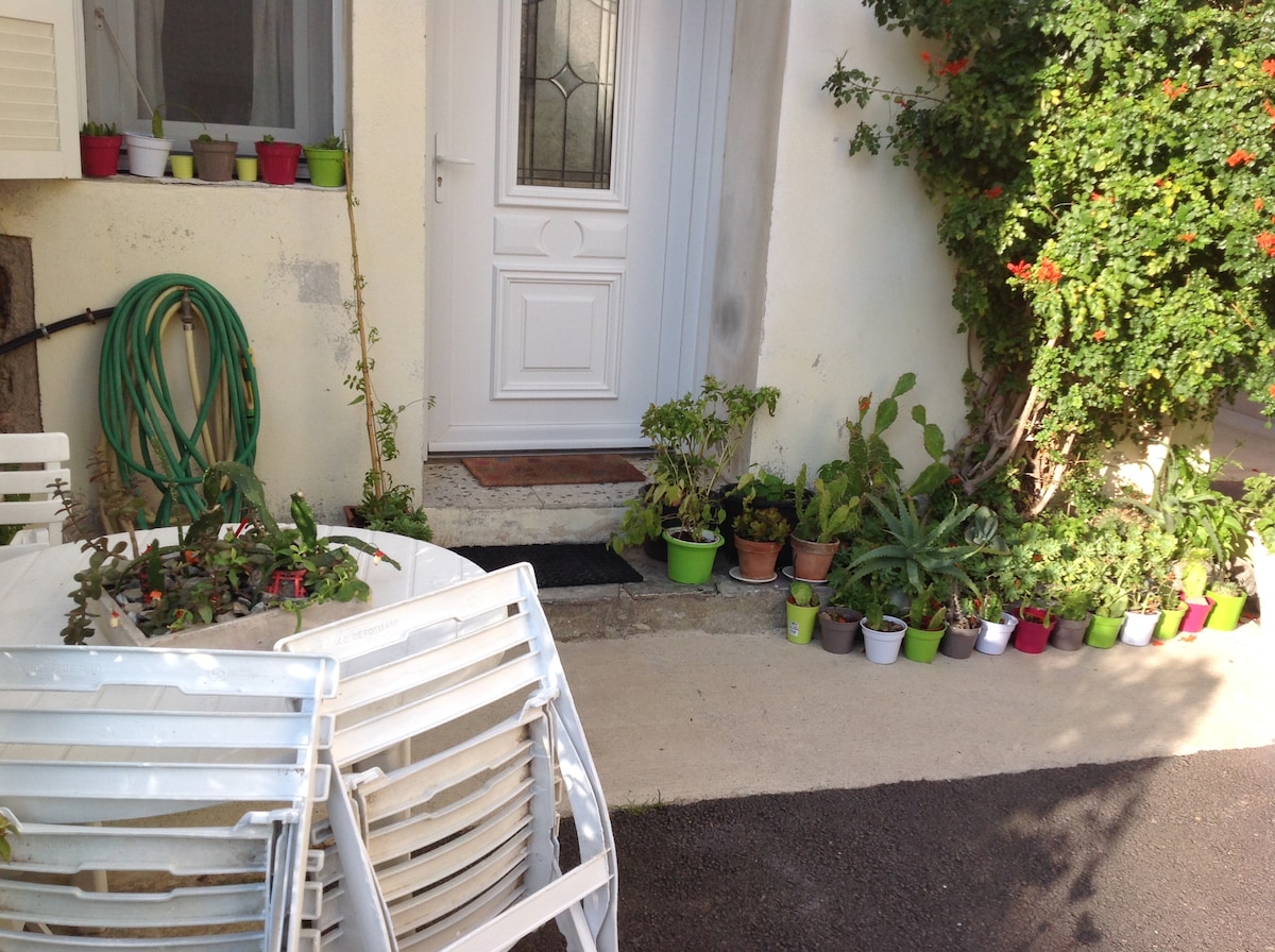 A welcoming entrance features a white door with a glass panel set into a light-colored wall. Colorful pots with various plants line the pathway, accompanied by stacked white chairs. A green garden hose is positioned beside the door, adding to the outdoor charm.
