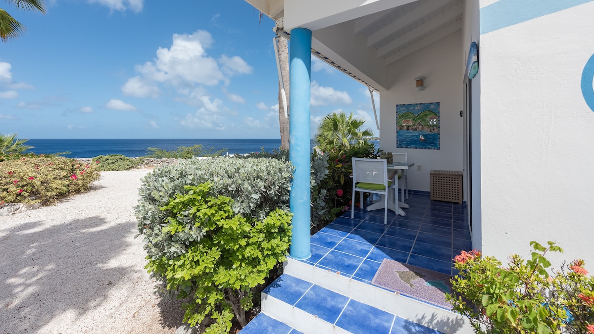 The entrance is framed by vibrant greenery and colorful flora, leading to a shaded patio area with a dining table and chairs. A blue-tiled floor extends towards the ocean, which is visible in the background, complemented by a bright sky and fluffy clouds.