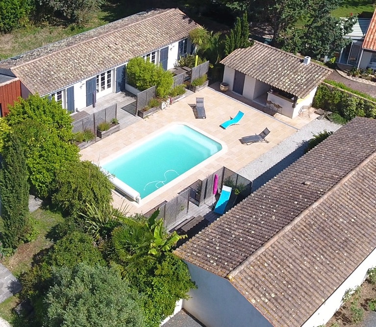 An aerial view showcases three adjacent cottages surrounded by greenery. A non-heated pool is centered in a tiled area with sun loungers and a barbecue space. Lush trees and shrubs provide natural framing, enhancing the tranquil atmosphere of the outdoor space.