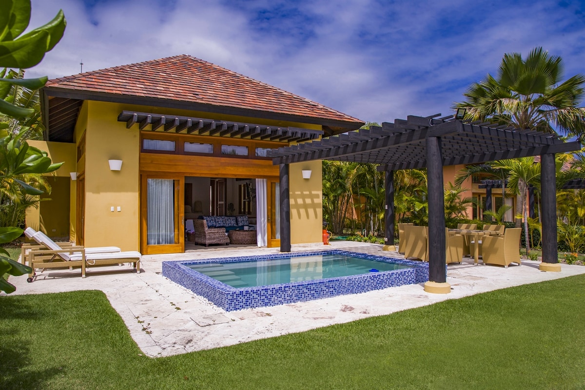 A private pool is surrounded by lush greenery, with a pergola providing shade over a dining area. The bungalow features large sliding doors that open to reveal a comfortable seating area inside, enhancing the connection to the outdoor environment.