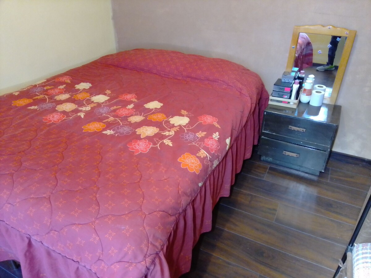 A spacious bed is dressed with a floral-patterned quilt in warm tones. A wooden bedside table is positioned nearby, featuring several stacked items and a small mirror. Dark wooden flooring complements the cozy ambiance of the sleeping area.