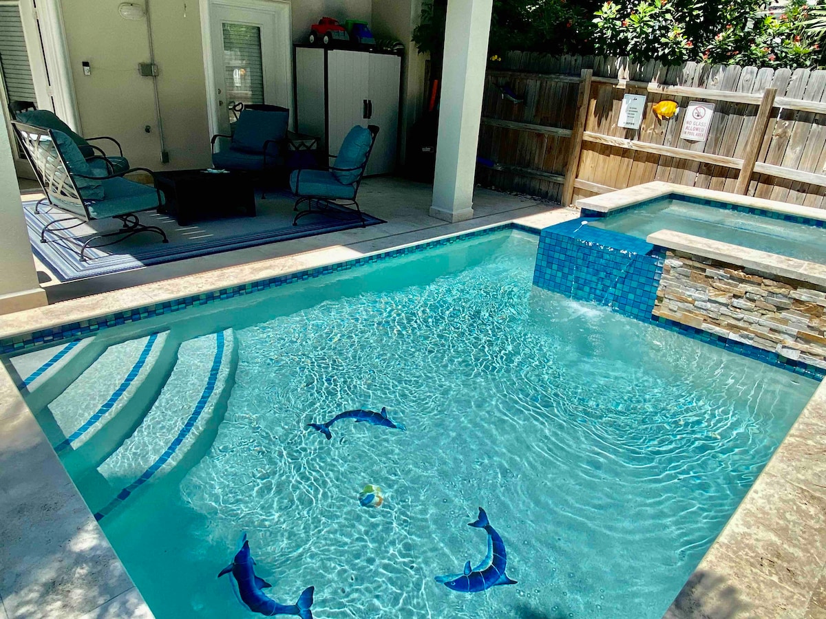 A small private heated pool features a shimmering blue surface with dolphin designs. Nearby, a seating area is arranged with two blue chairs and a table, providing a space for relaxation. Sunlight reflects off the water, enhancing the tranquil setting.