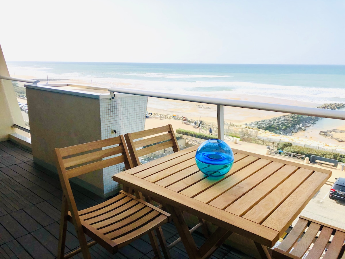 A terrace features a wooden table paired with two folding chairs, positioned to overlook the ocean. A decorative blue glass orb is placed at the center of the table, while the expansive sea and sandy beach can be seen in the background.