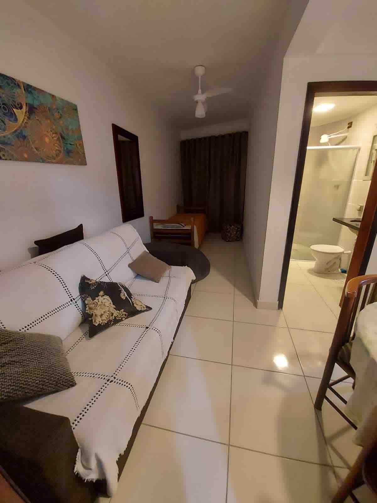 A simple seating area features a light-colored sofa adorned with decorative cushions, positioned against a neutral wall. A tasteful wall art piece adds visual interest. A glimpse of a hallway leads to a bathroom area, which is separated by a partially transparent door.