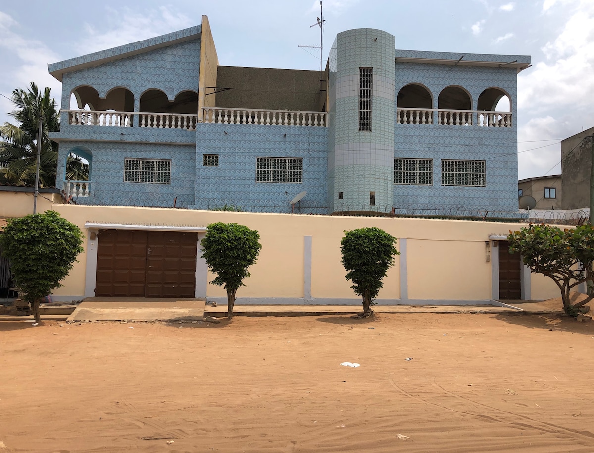 The exterior of a two-story building is displayed, showcasing a distinctive blue façade with intricate tile patterns and multiple arched windows. Two garage doors are visible on the ground level, complemented by well-maintained trees and a sandy open space in the foreground.