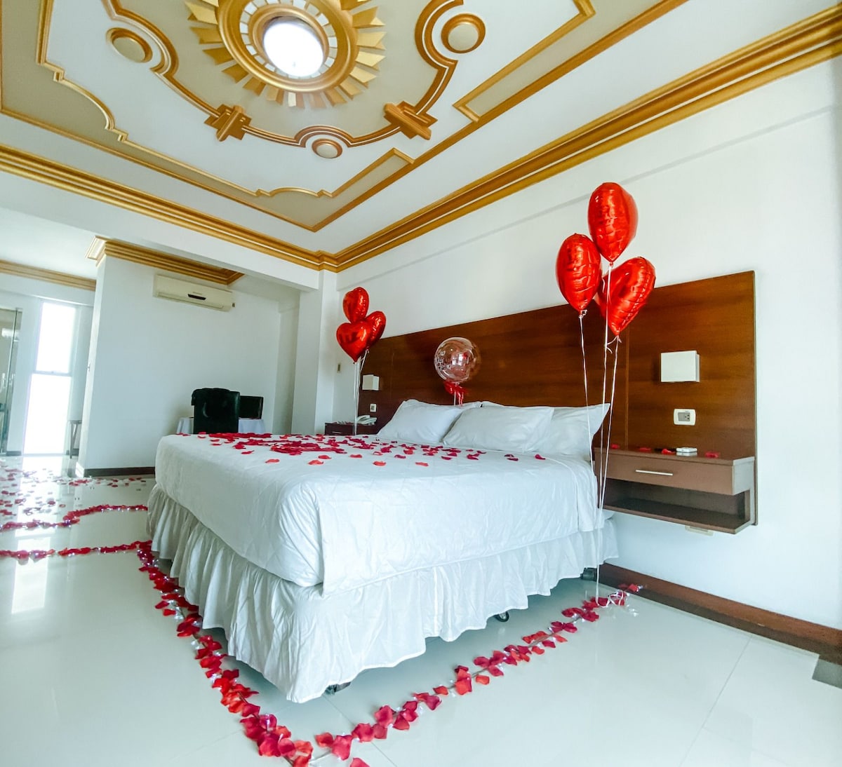 A bright bedroom features a spacious bed adorned with crisp white linens. The floor is decorated with rose petals, and red heart-shaped balloons are placed around the room. Elegant crown molding accents the ceiling, while natural light enters through a nearby window.