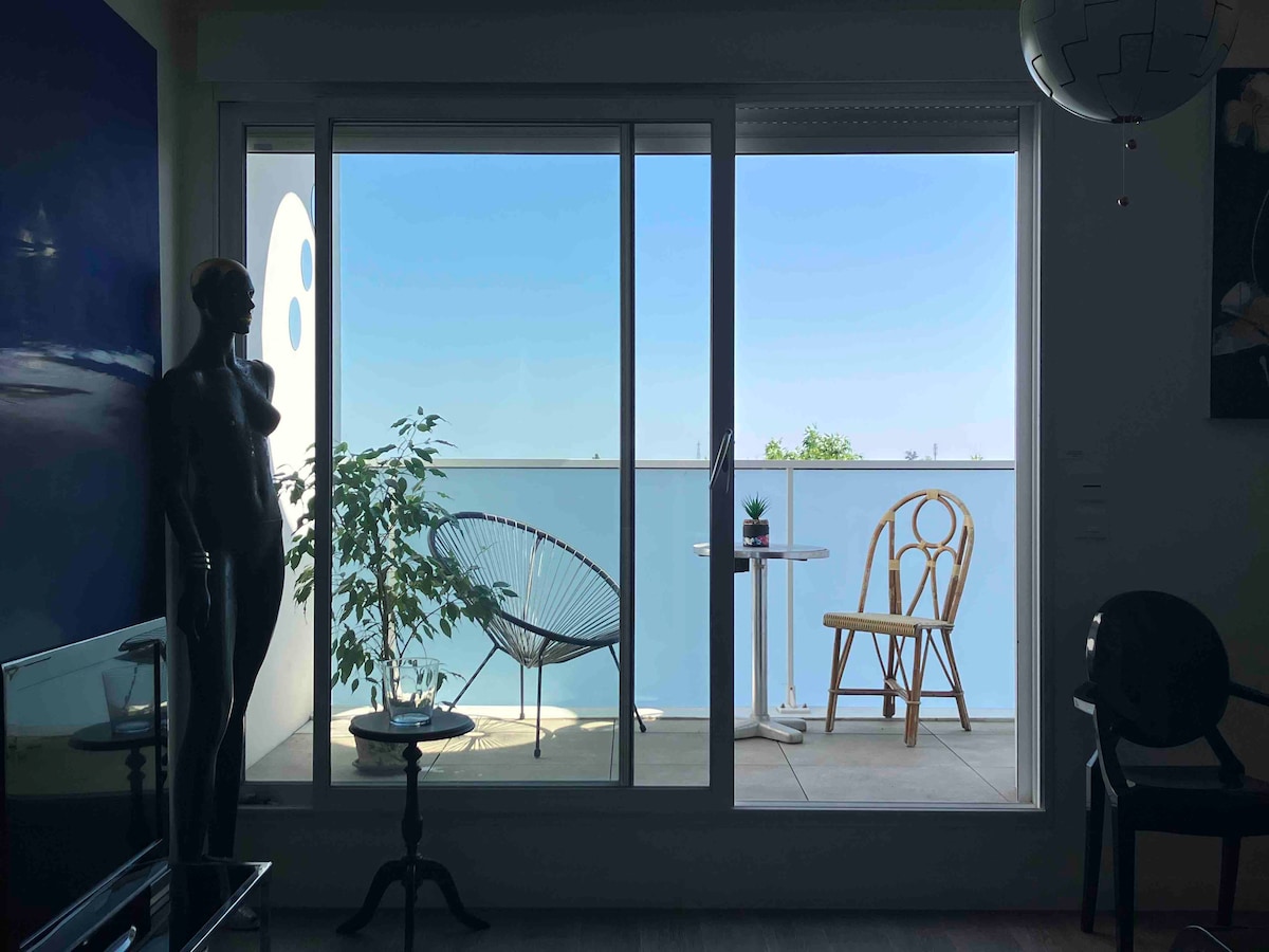 A view of a balcony is framed by large glass doors, showcasing a bright blue sky. A single chair and a small table are placed on the balcony, with a potted plant adding a touch of greenery. A modern sculpture and a chair are visible in the foreground.