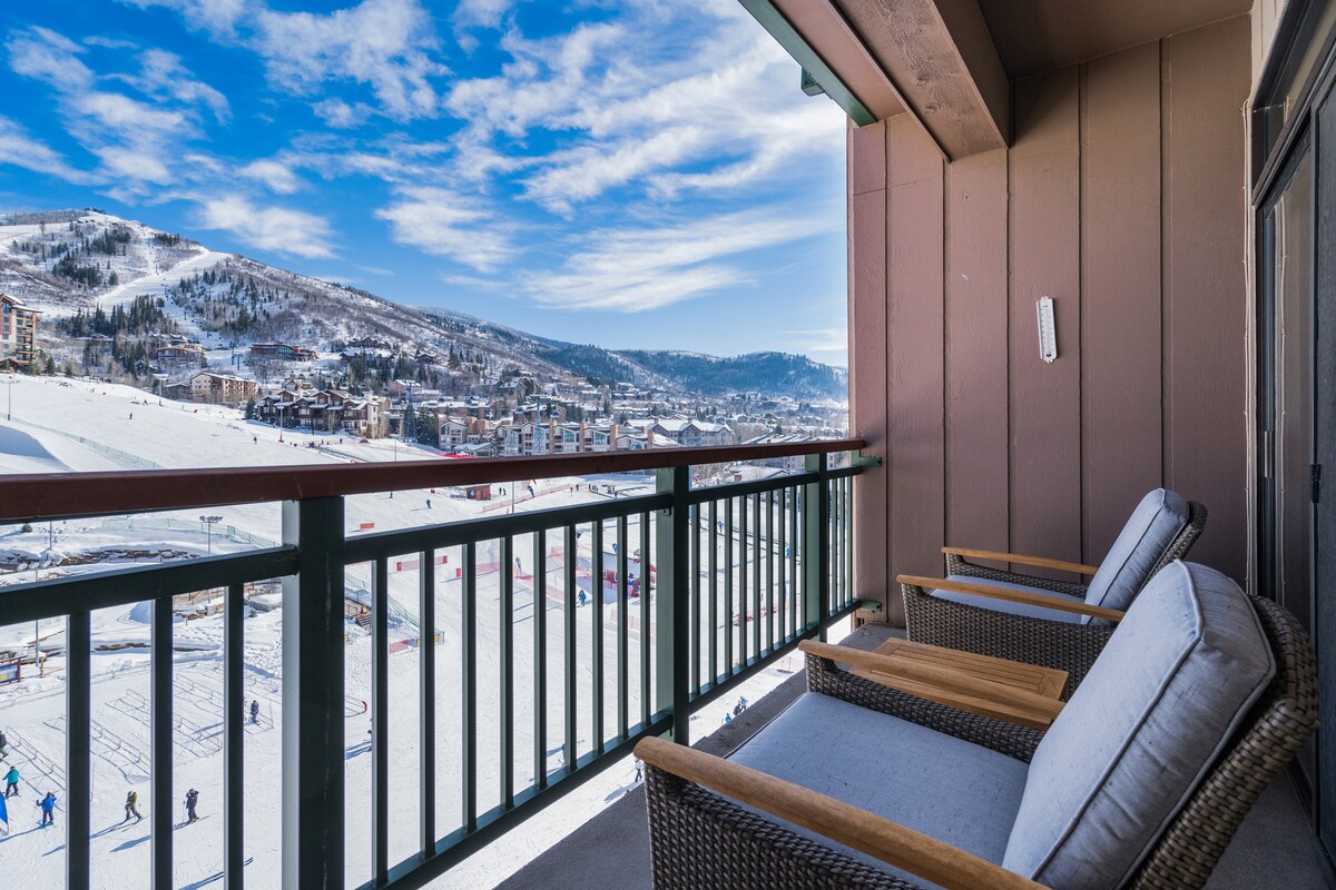 A private balcony features two armchairs facing expansive views of the ski slopes and surrounding mountains, under a bright sky. The railing is designed in a muted green, complementing the warm tones of the wooden walls.