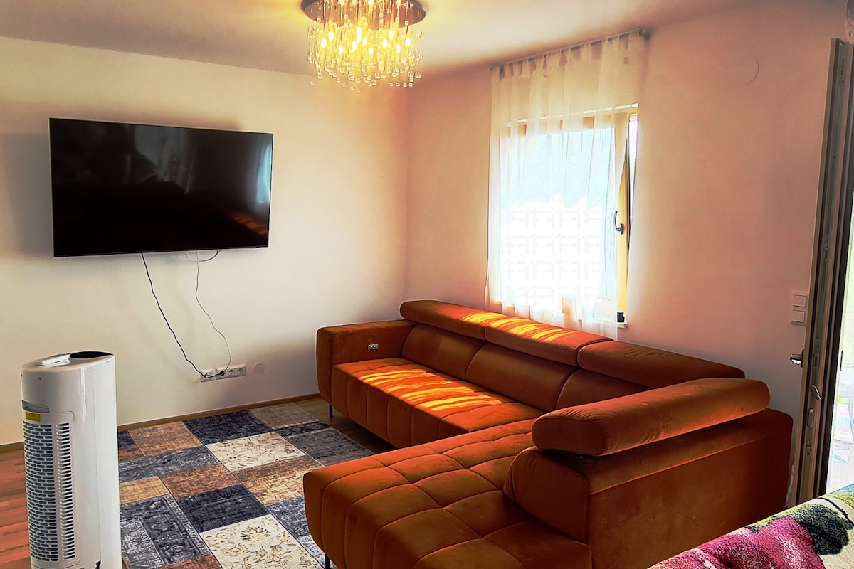 A modern living area features a large orange sectional sofa and a flat-screen television mounted on the wall. Natural light fills the space through a window adorned with sheer curtains. A patterned area rug adds texture to the floor, and a portable heater stands nearby.