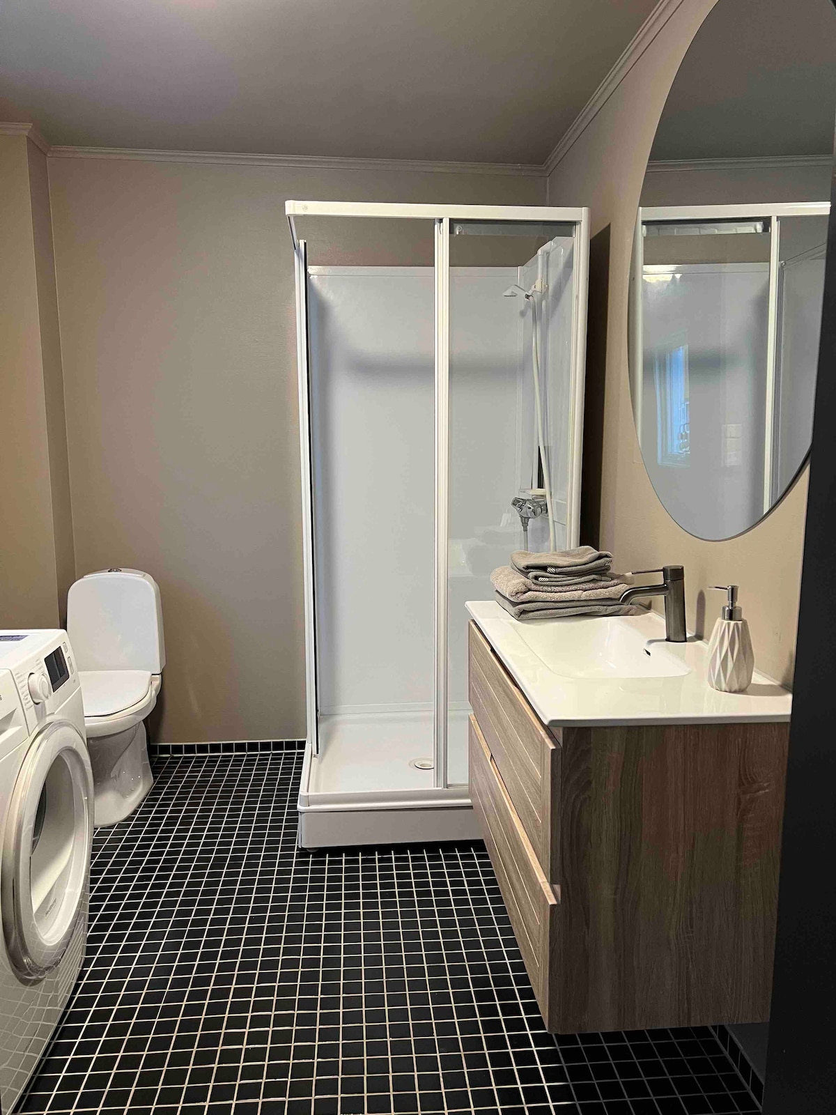 A compact bathroom features a standing shower with a glass door and modern sink cabinet. A round mirror reflects the space, while neatly stacked towels are placed on the counter. A washing machine is positioned beside the toilet, all set against dark tiled flooring.