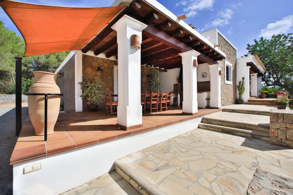 An outdoor dining area is seen, featuring a large table surrounded by wooden chairs. A traditional clay pot is placed on the edge of the stone patio. Pillars support a shaded terrace, while stone steps lead to the entrance of the home amidst a natural landscape.