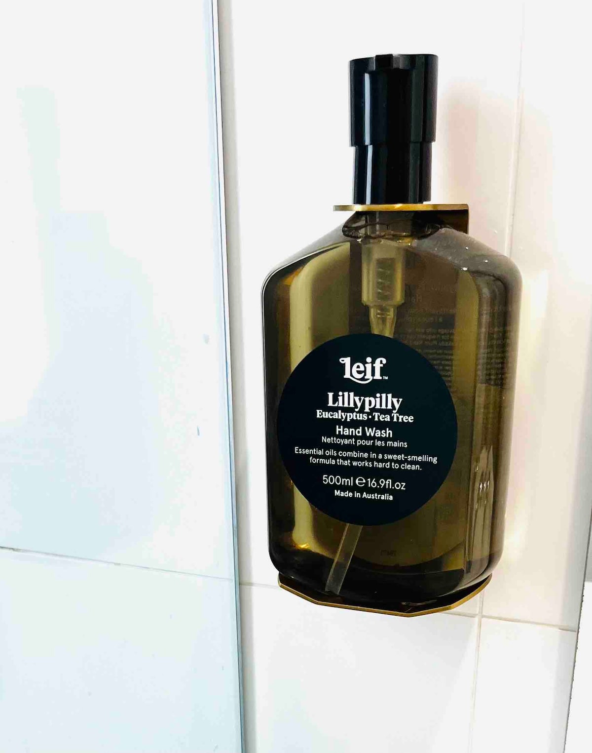 A green amber hand wash bottle is affixed to a white tiled wall. The label indicates it contains Lillypilly Eucalyptus Tea Tree formula, emphasizing natural ingredients. The sleek design and label clarity provide an organized and thoughtful touch to the apartment's amenities.