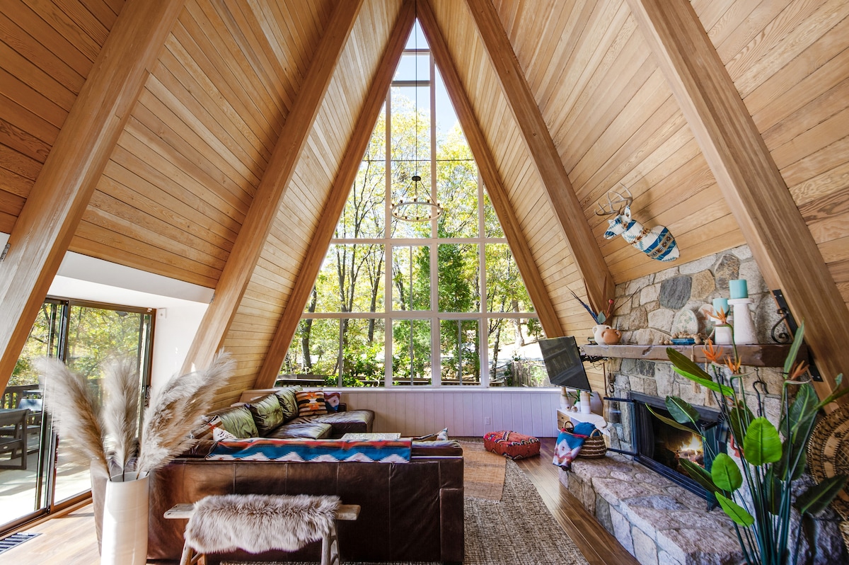 The spacious great room features a high A-frame wooden ceiling and large glass windows that provide abundant natural light and views of the surrounding trees. A fireplace with faux logs is positioned against a stone wall, accompanied by a comfortable seating area adorned with colorful throw pillows.