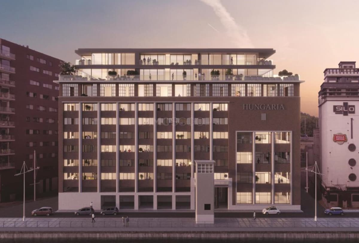 The recently renovated Hungaria building showcases a modern architectural design with multiple stories, large glass windows, and a rooftop terrace. The structure is situated along a waterfront, surrounded by other urban settings. Warm light is visible through the windows, highlighting the building's inviting presence.