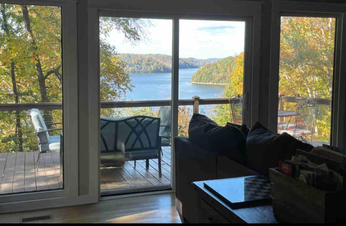 Large sliding glass doors open to a scenic view of the lake, framed by autumn foliage. A cozy seating area is visible on the deck, complemented by two chairs. Soft natural light fills the interior space, creating an inviting atmosphere.