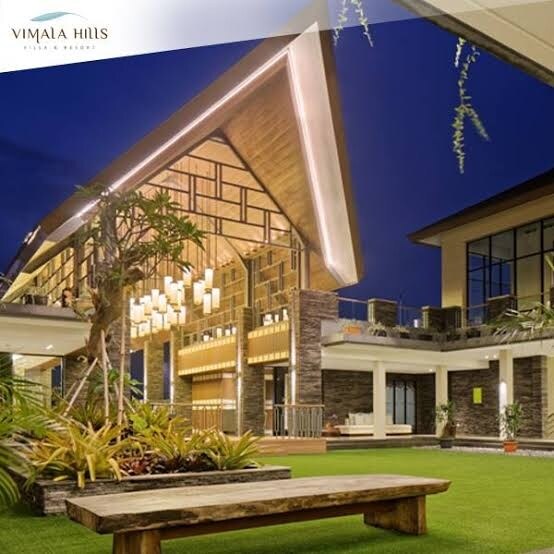 An inviting outdoor area features a spacious wooden bench surrounded by lush greenery and well-maintained landscaping. The modern architecture is highlighted by a striking roof design and warm lighting, creating a serene environment. Large windows offer glimpses into the villa's interior.