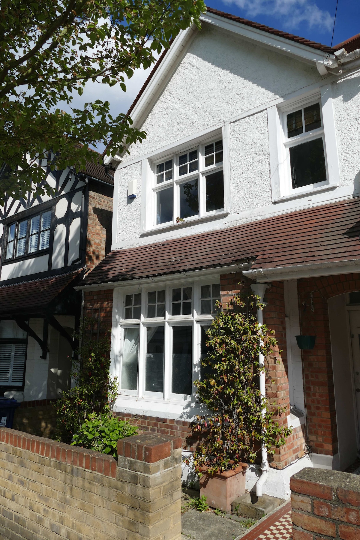 A charming white house is shown, featuring multiple windows framed by green foliage. The brick façade is complemented by a patterned walkway leading to the entrance. Sunlight filters through the leaves, creating a serene and inviting appearance.